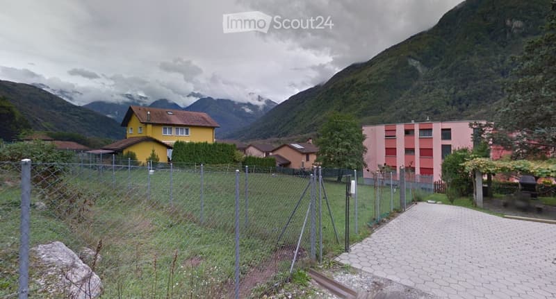 A fenced garden area with a paved pathway leading to a residential complex, surrounded by trees and mountains in the background.