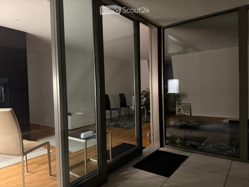 Modern interior with large glass sliding doors leading to a balcony. Visible furniture includes a white chair, small table, and a lamp in the adjacent room.