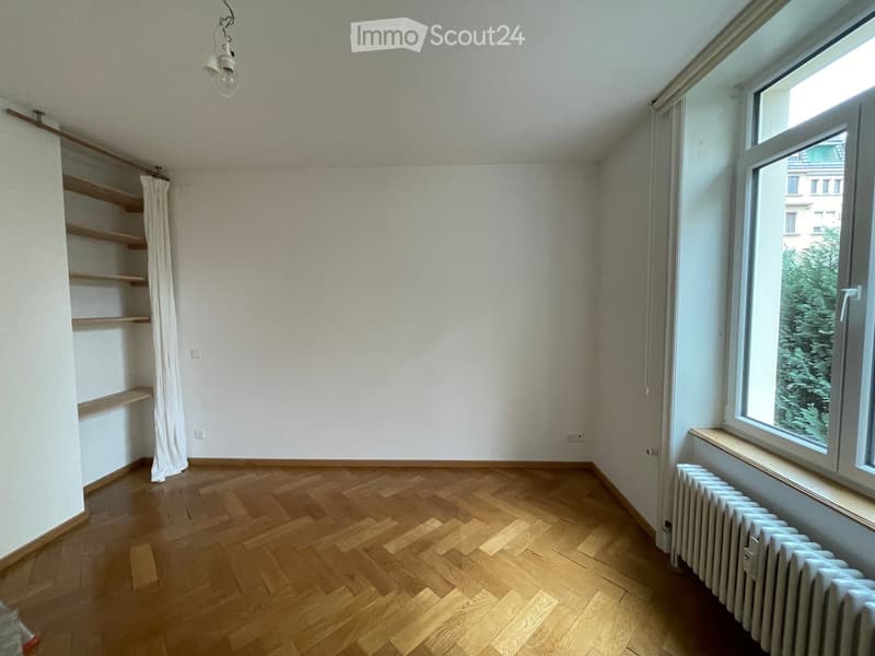 Empty room with wooden flooring, white walls, a window with a view, a radiator, and built-in wooden shelves.
