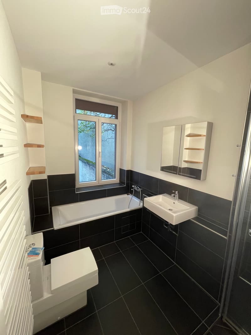 Modern bathroom with black tiles, white fixtures, a bathtub, sink, toilet, mirror, and built-in shelves.