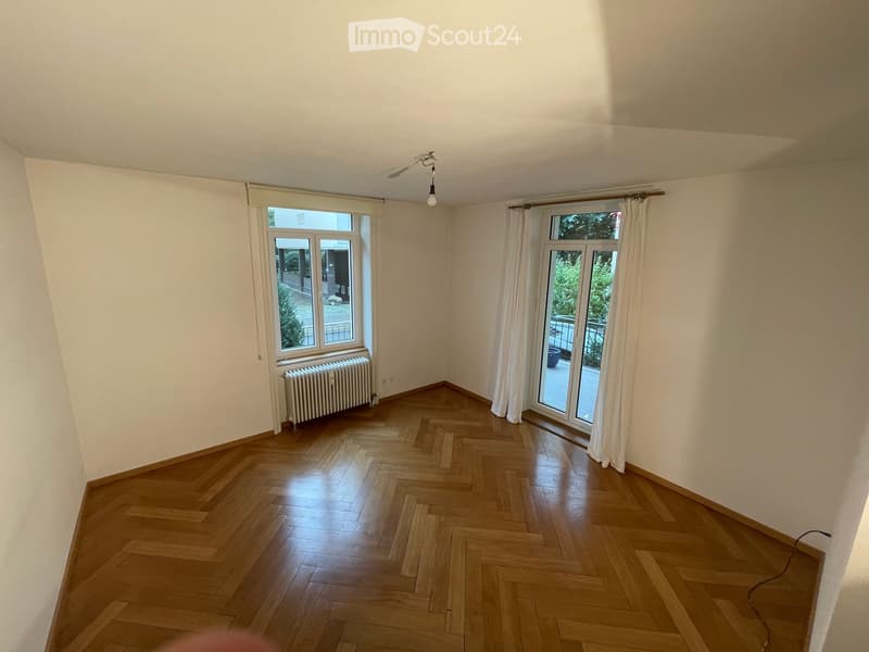 Empty room with wooden herringbone flooring, white walls, large windows with white curtains, radiator, and a ceiling light fixture.