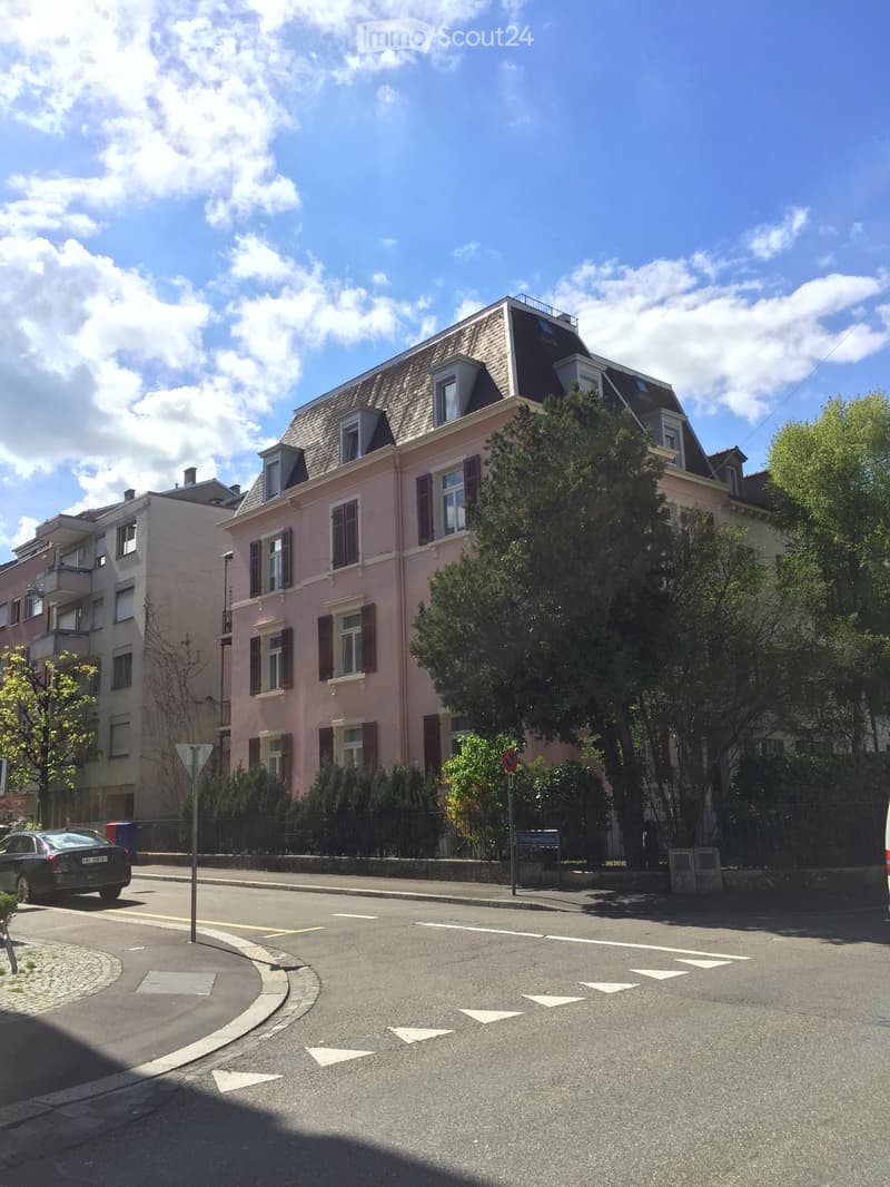 Pink residential building with multiple windows and shutters, surrounded by trees and greenery, with a street and parked car visible.