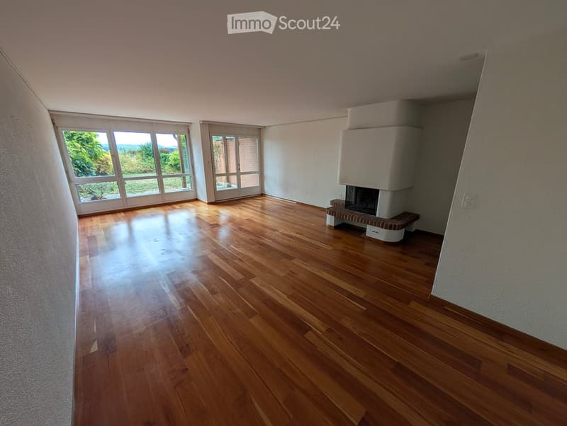 Empty room with wooden flooring, large windows, and a fireplace with a bench.