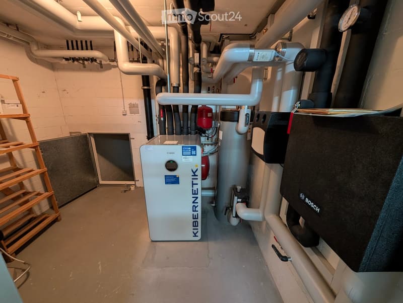 Utility area with heating and water systems, including a Bosch and Kibernetik unit, surrounded by white pipes and a wooden shelf on the left.