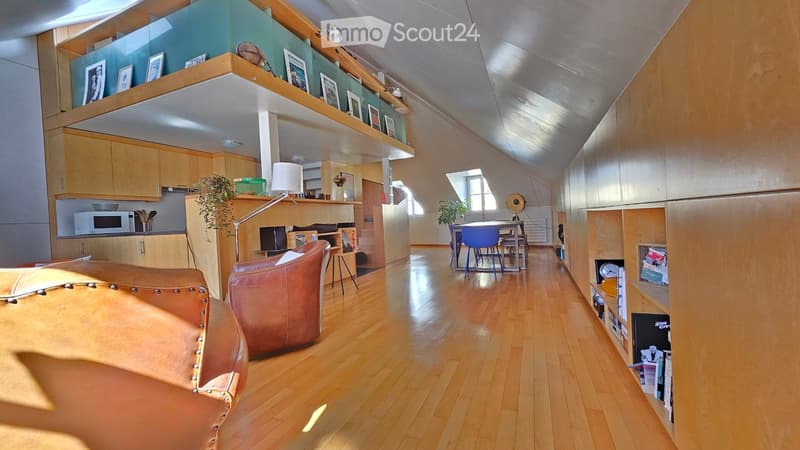 Open-plan living and dining area with wooden flooring, modern furniture, and built-in shelving. Features a leather sofa, dining table with chairs, microwave, and decorative items on shelves.