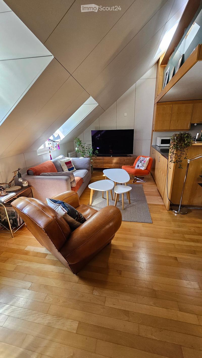 Modern living room with wooden flooring, a grey sofa, leather armchair, coffee table, small stools, a television, and a small kitchen area with cabinets and a microwave.