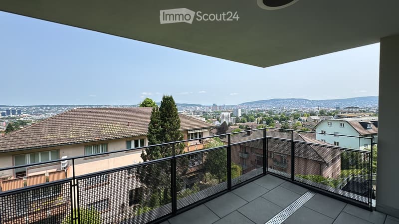 Balcony with a metal railing, overlooking a cityscape with houses, trees, and distant mountains.