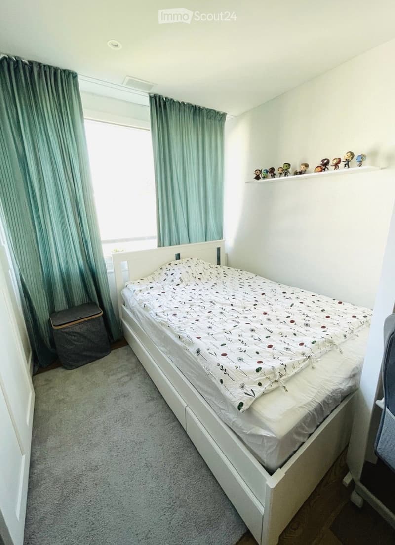 A tidy bedroom with a white bed, green curtains, a shelf with figurines, and a grey trash bin.