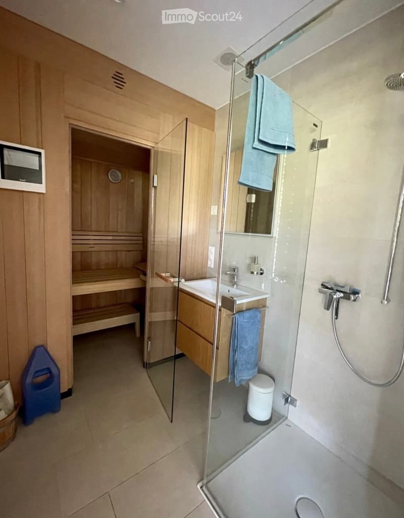 Modern bathroom with a glass-enclosed shower, double sink, and sauna area. Features include a showerhead, towel rack, and a small trash can.