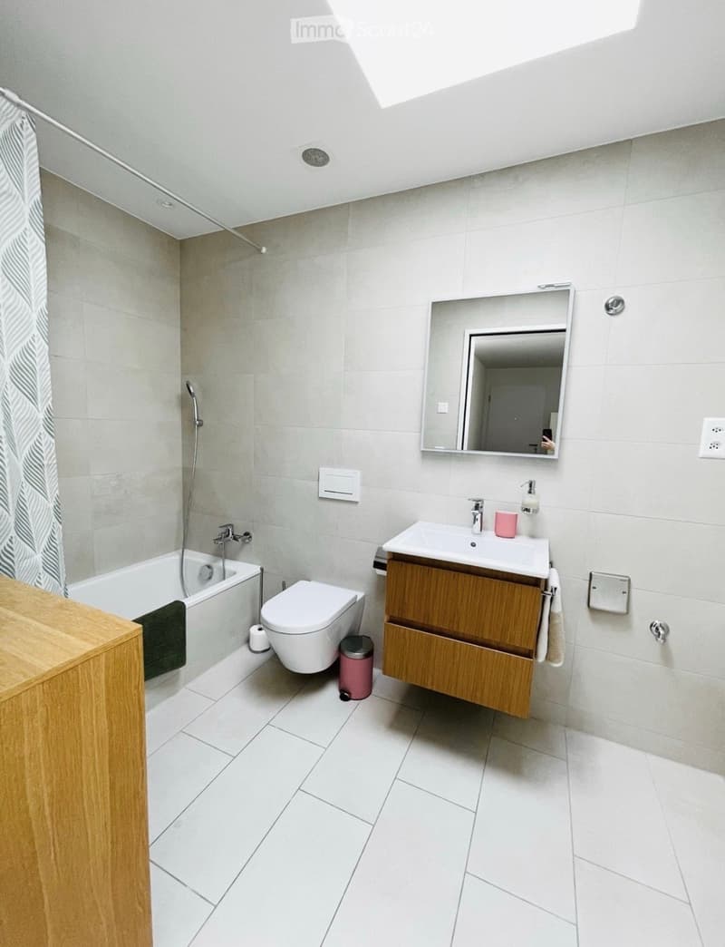 Modern bathroom with white tiles, a bathtub, toilet, sink with wooden cabinet, mirror, and a shower curtain.