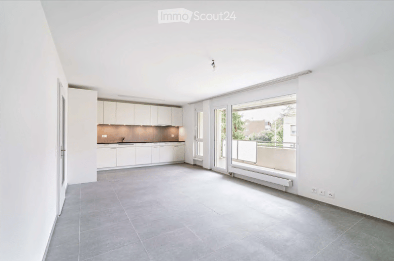 Open-plan space with a modern kitchen area featuring white cabinets and a sink, connected to a living area with large windows and a balcony access. The floor is tiled, and the walls are painted white.