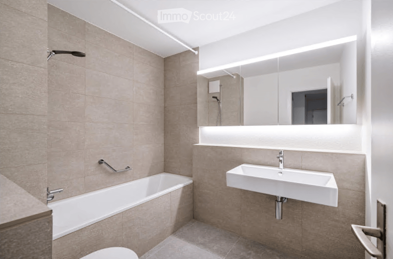 Modern bathroom with a bathtub, sink, and large mirror with integrated lighting. Beige tiles and sleek fixtures.