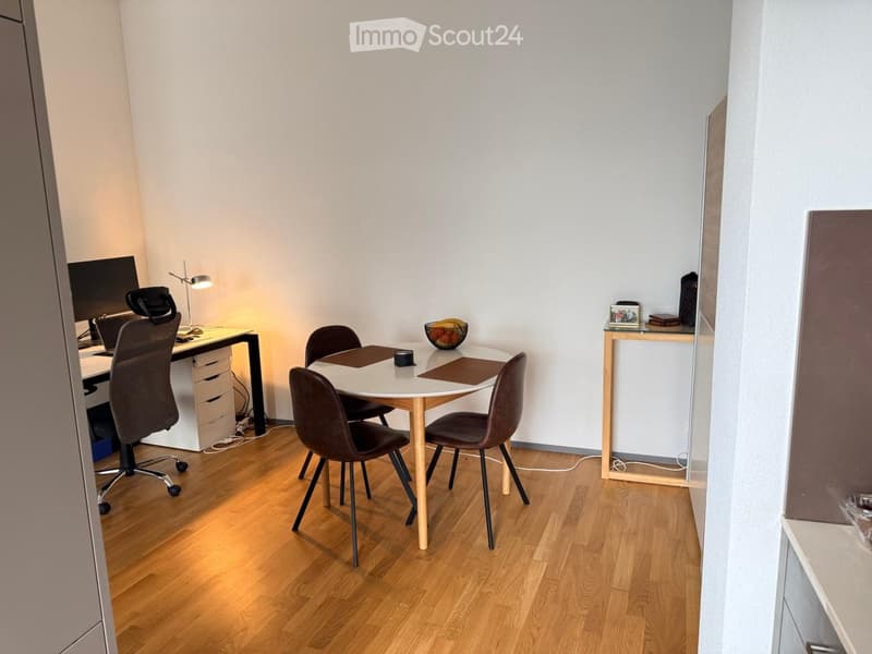 A modern, well-lit room with a wooden floor, featuring a desk with a chair, a round dining table with four chairs, and a small shelf with decorative items.