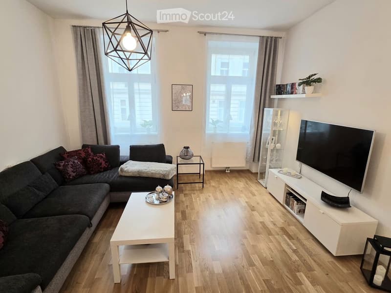 Modern living room with wooden flooring, large windows, grey sectional sofa, white coffee table, TV stand, side table, wall shelves, and a geometric hanging light fixture.