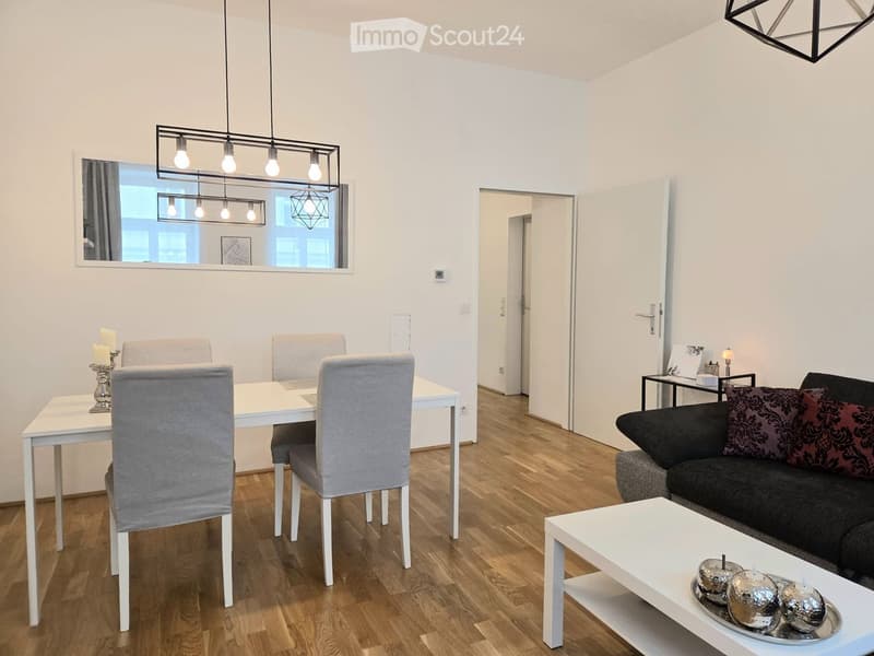 Modern living and dining area with light wood flooring, white walls, and contemporary furniture including a sofa, coffee table, dining table, and chairs.