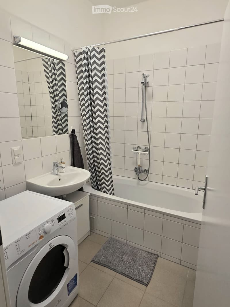 Modern bathroom with white tiles, a sink, bathtub with shower, washing machine, and a towel on the wall.