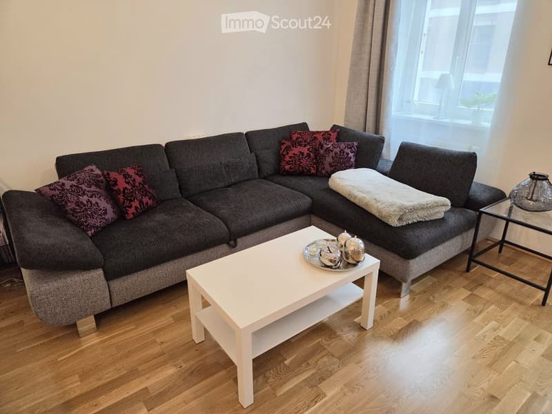 Modern living room with a grey sectional sofa, white coffee table, decorative tray with candles, and a glass table lamp. The room features wooden flooring and large windows with sheer curtains.
