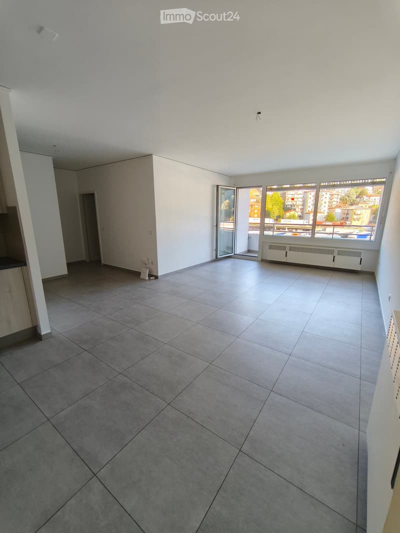 Empty room with tiled floor, white walls, large windows, and an open sliding door leading to a balcony.