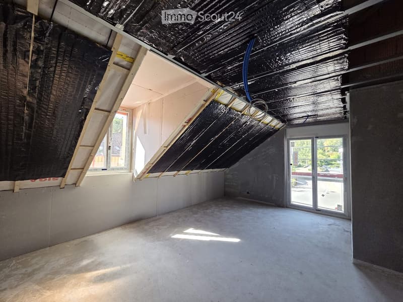 Empty attic space with exposed wooden beams, insulation, and a large window letting in natural light. The floor is concrete, and the walls are unfinished.