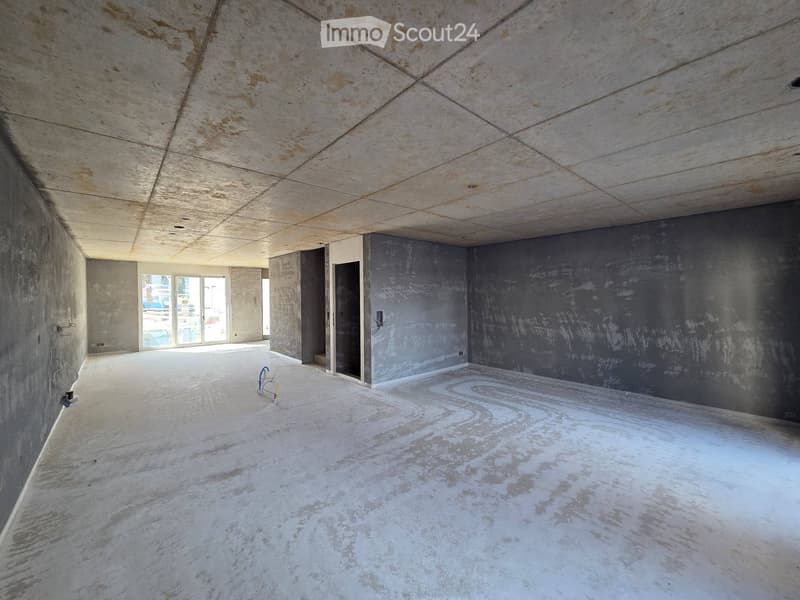Empty room with bare concrete walls and ceiling, unfinished surfaces, and a large window letting in natural light.