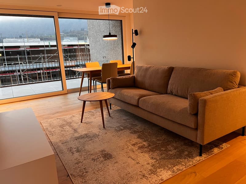 Modern living room with a brown sofa, coffee table, dining table, and chairs, large windows with a view, and wooden flooring.