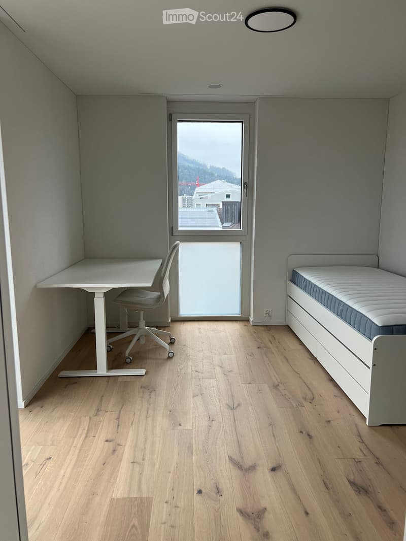 A small, modern room with light wooden flooring, a white desk with a chair, and a built-in bed with a mattress and storage unit.