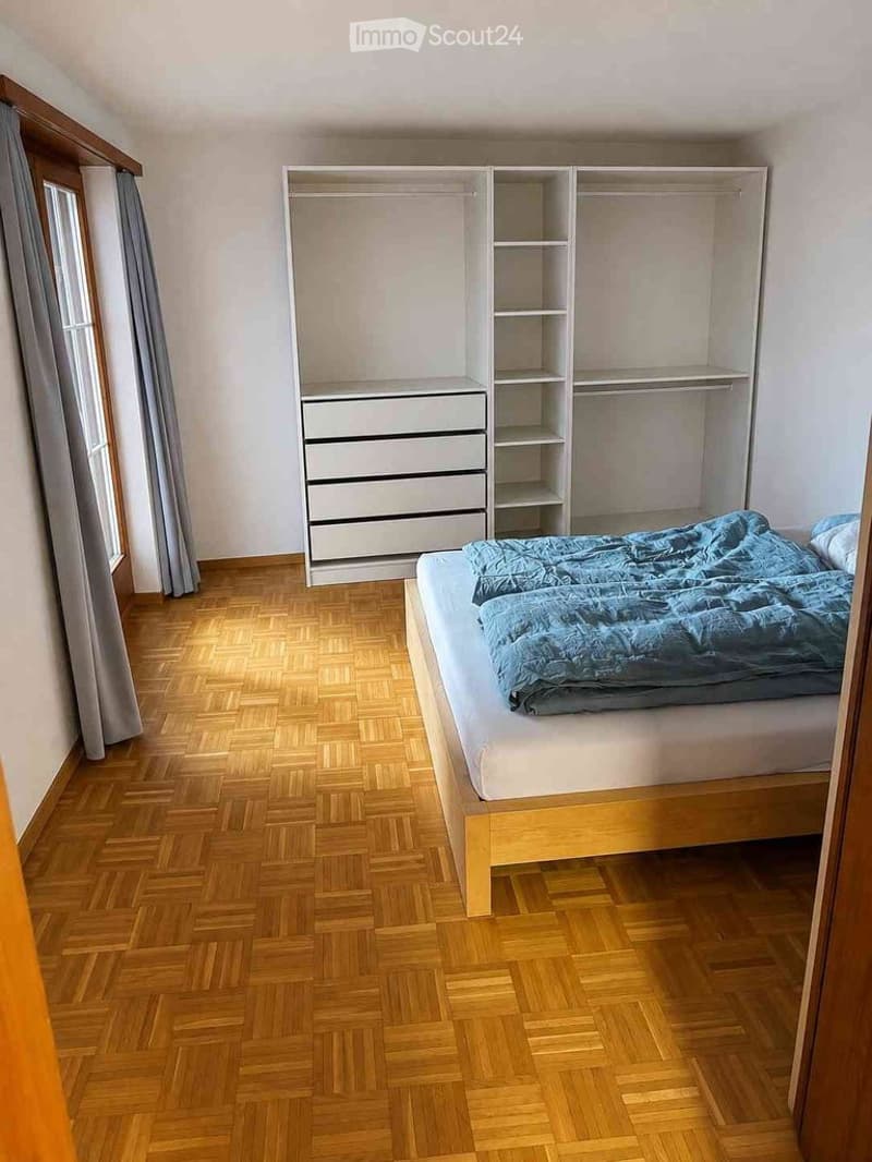 Bedroom with wooden herringbone flooring, a single bed with blue bedding, a built-in wardrobe with shelves and drawers, and a window with grey curtains.