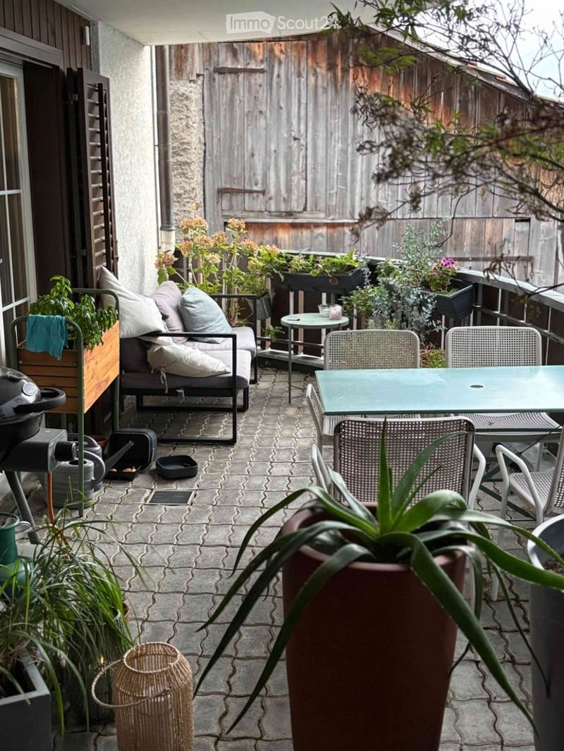 A cozy outdoor balcony with a seating area, small table, chairs, and various potted plants. The space features a brick floor and wooden fencing.