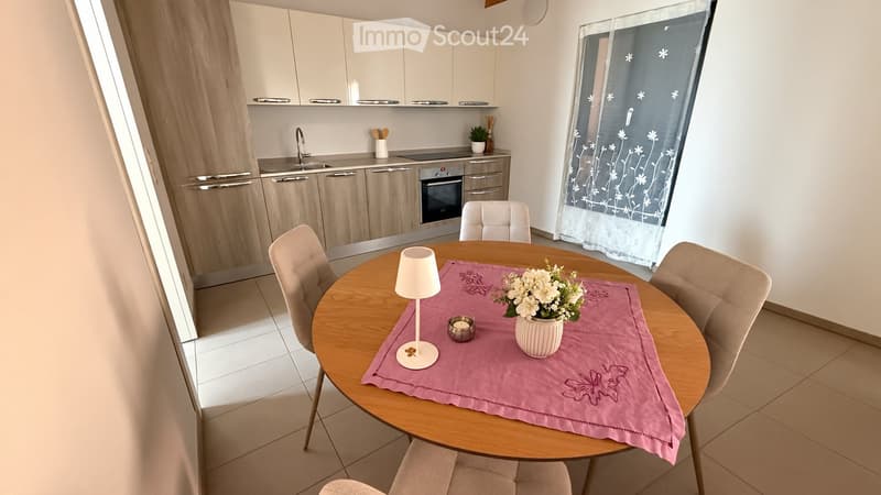 A modern, well-lit dining area with a round wooden table, four beige chairs, a white lamp, a vase with flowers, and a pink table runner. The adjacent kitchen features light wood cabinets, stainless steel appliances, and a sink.