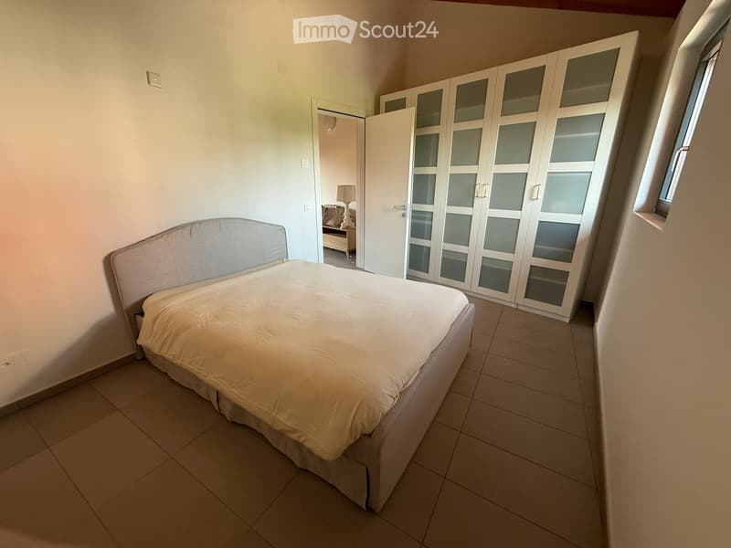A well-lit bedroom with a neatly made bed, a large wardrobe with glass doors, and a small lamp visible in the adjacent room.