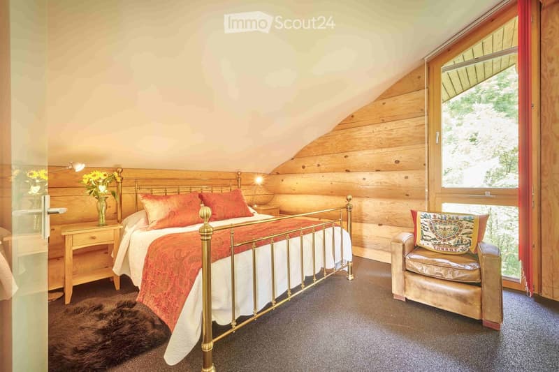 Furnished attic bedroom with wooden wall and ceiling, double bed, nightstand, lamp, armchair, rug, carpeted floor, and a window with view of the forest.