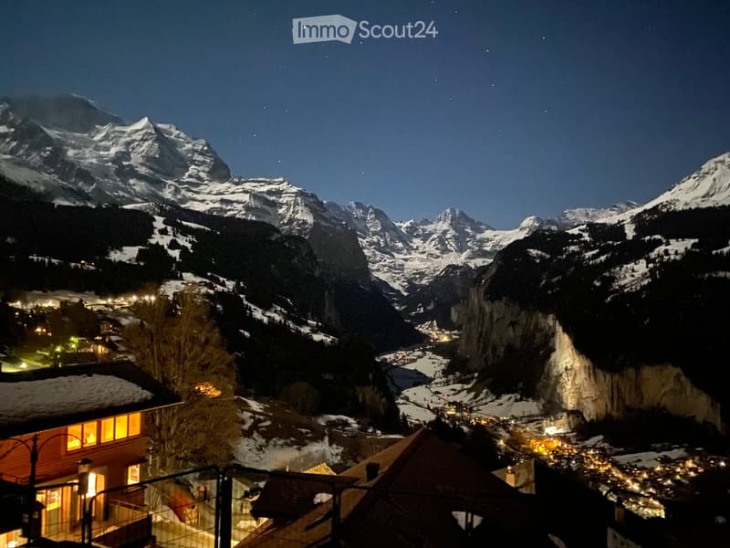 Wooden house on hilltop with mountains in the background, and a river valley with lights below