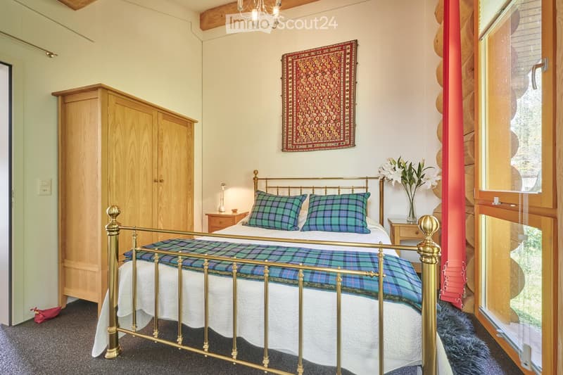 a bedroom with a brass bed, wooden wardrobe, wooden nightstands, carpeted floor, wall lamp, and flowers in a vase on a nightstand, and a rug hanging on the wall