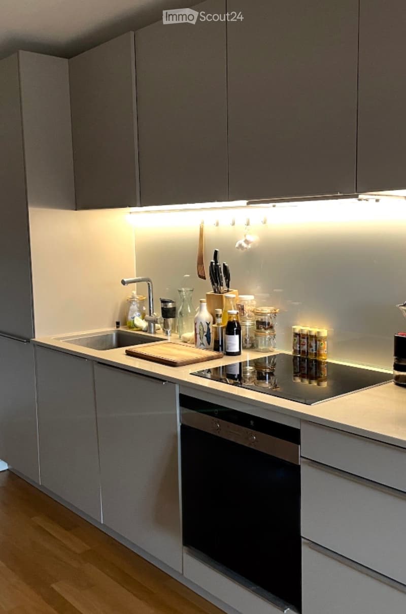 Modern kitchen with grey cabinets, stainless steel appliances, and a sleek countertop. Features include a sink, faucet, cutting board, blender, oven, and various kitchen items on the counter.
