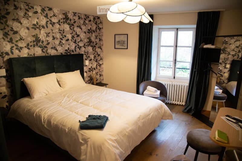 Cozy bedroom with a large double bed, nightstand, chair, small table, and mirror. The room features patterned wallpaper, wooden flooring, and a window with dark curtains.