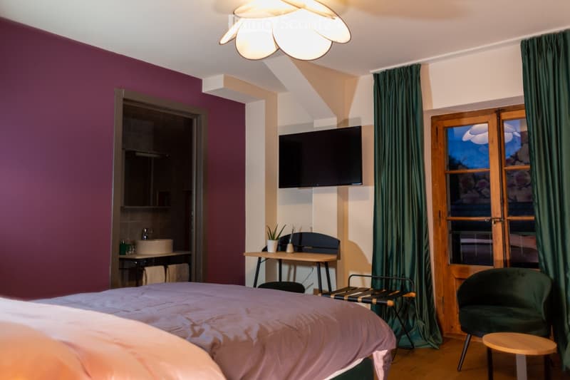 Cozy bedroom with a bed, a chair, a small table, a TV screen, a sink, and a curtained window.