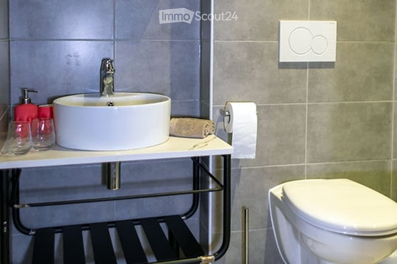 Modern bathroom with a white sink, chrome faucet, toilet, and a towel on the counter. There are two glasses and a soap dispenser on the counter as well.