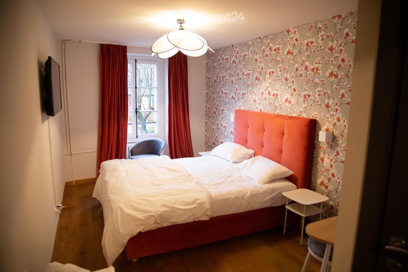 Cozy bedroom with a double bed, red headboard, white bedding, a small table, a chair, and a window with red curtains. The room features a floral wallpaper and a modern pendant light.