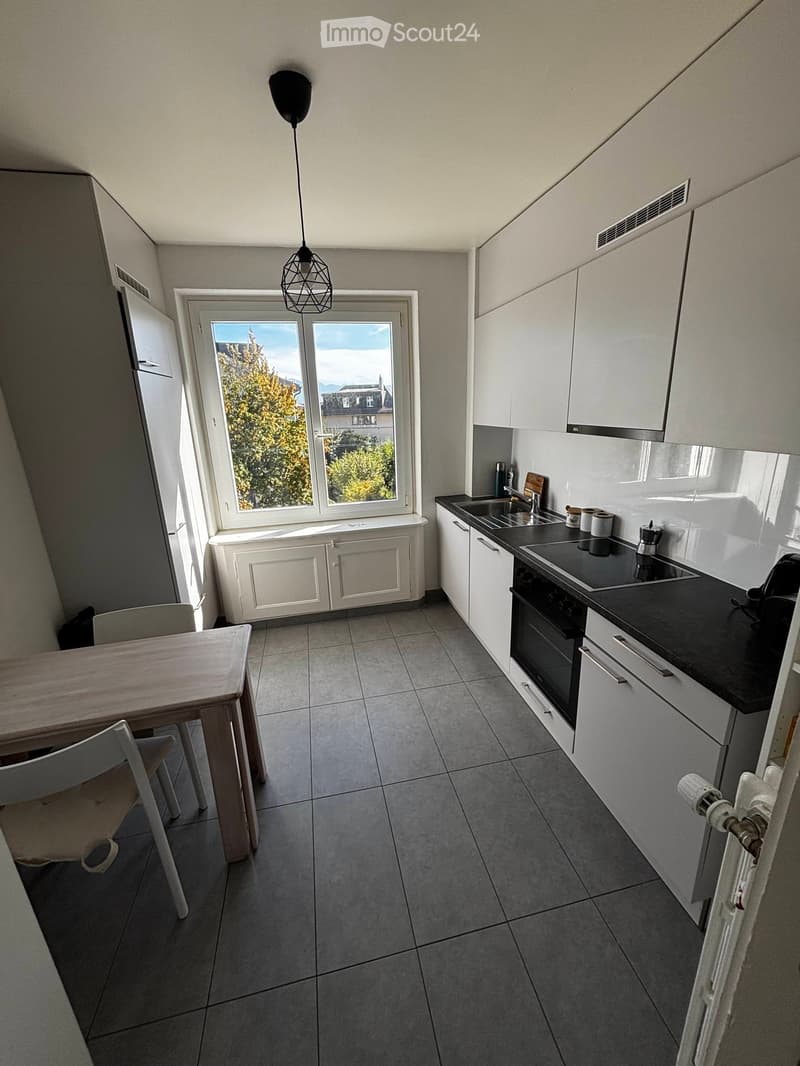 Modern kitchen with white cabinets, black countertops, a built-in oven, sink, and a small dining area with a table and chairs. The room has large windows offering a view of trees and buildings outside.