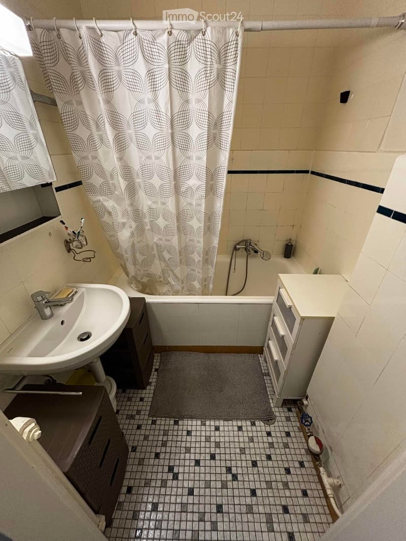 Compact bathroom with a sink, bathtub, shower curtain, and small cabinet. Features tiled walls and floor with a grey bathmat in front of the tub.