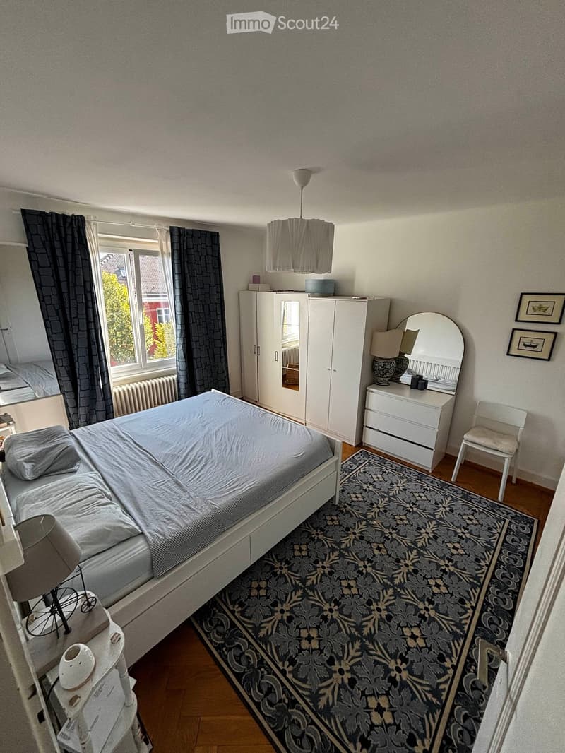Cozy bedroom with a double bed, white wardrobe, vanity with mirror, nightstand, and a patterned rug.