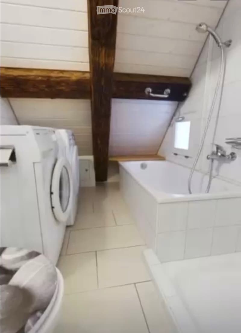 Modern bathroom with a bathtub, shower, and washing machine. The space features white tiled walls and floor, with a wooden accent wall and a built-in shelf.