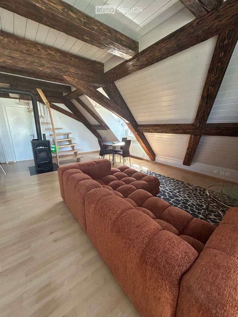 Attic living space with exposed wooden beams, a red tufted sofa, a glass coffee table, a dining area with chairs, and a fireplace.