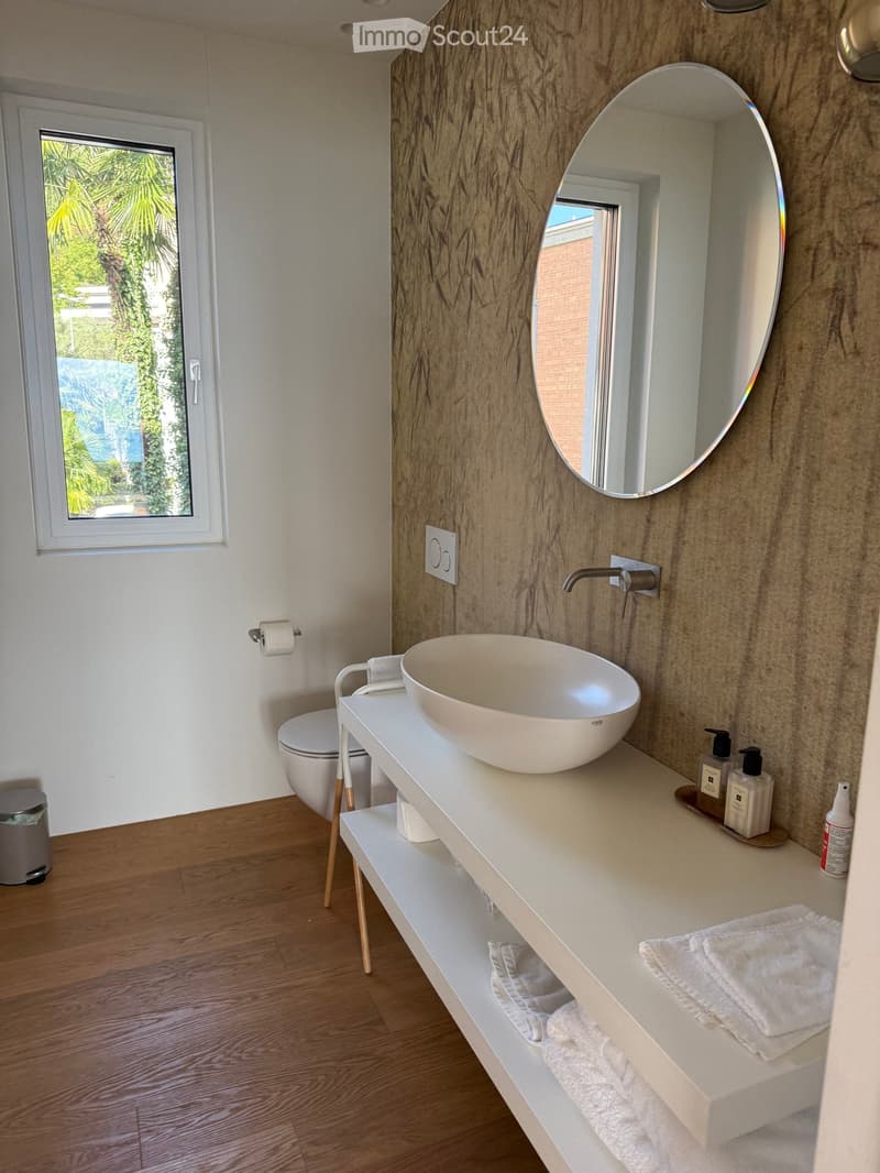 Modern bathroom with a white sink, round mirror, wooden floor, and a view of greenery through a window.