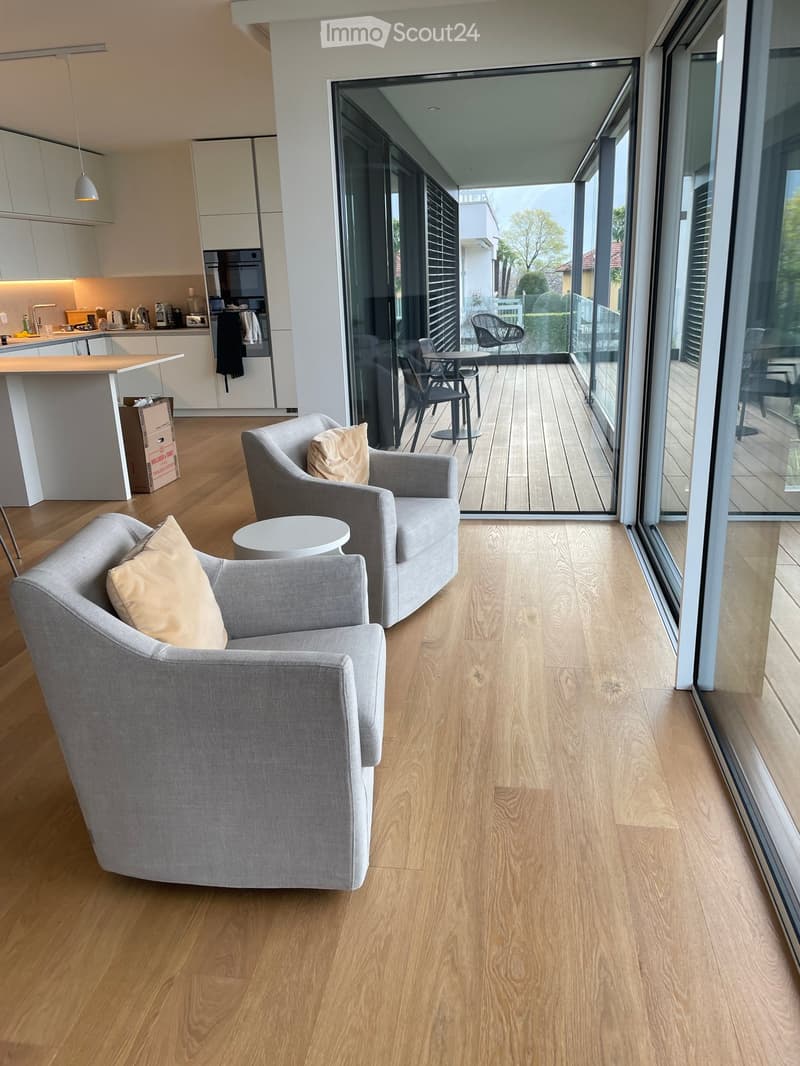 Modern living room with light wooden flooring, grey sofa, coffee table, and a view of a balcony with outdoor seating.