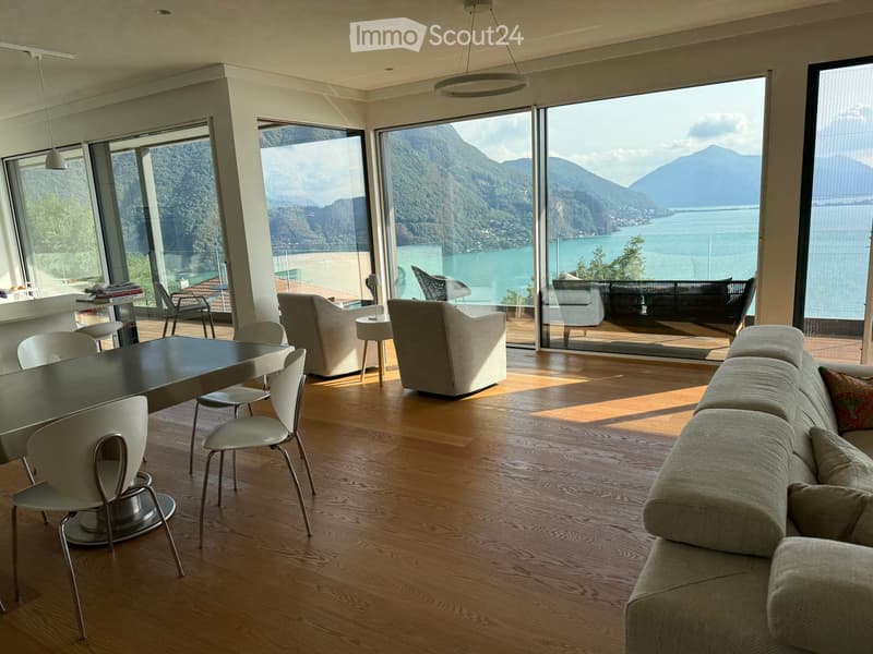 Modern living room with large windows offering a scenic mountain and lake view. Features a sectional sofa, coffee table, dining table with chairs, and several lamps.