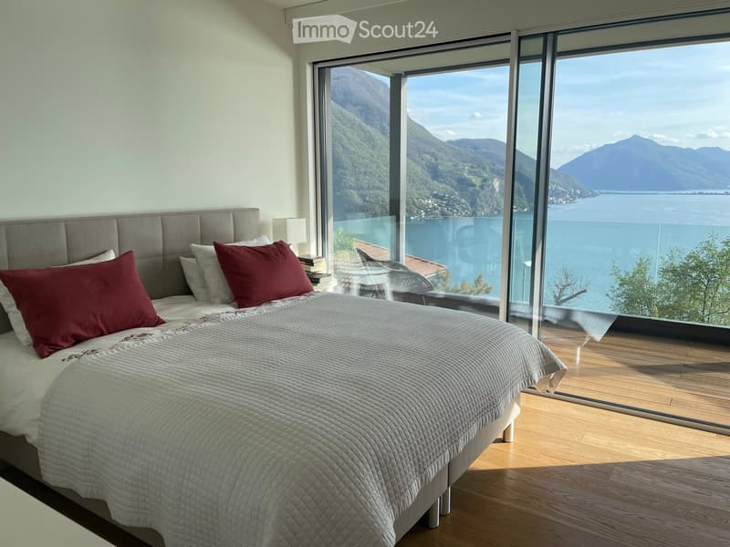 Modern bedroom with large windows offering a scenic mountain and lake view, featuring a grey upholstered headboard, a quilted bedspread, and red accent pillows.