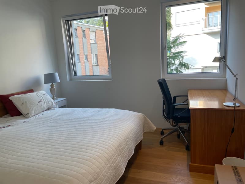 Cozy bedroom with a neatly made bed, a wooden desk with a chair, and a nightstand with a lamp. Large windows offer a view of the building outside.