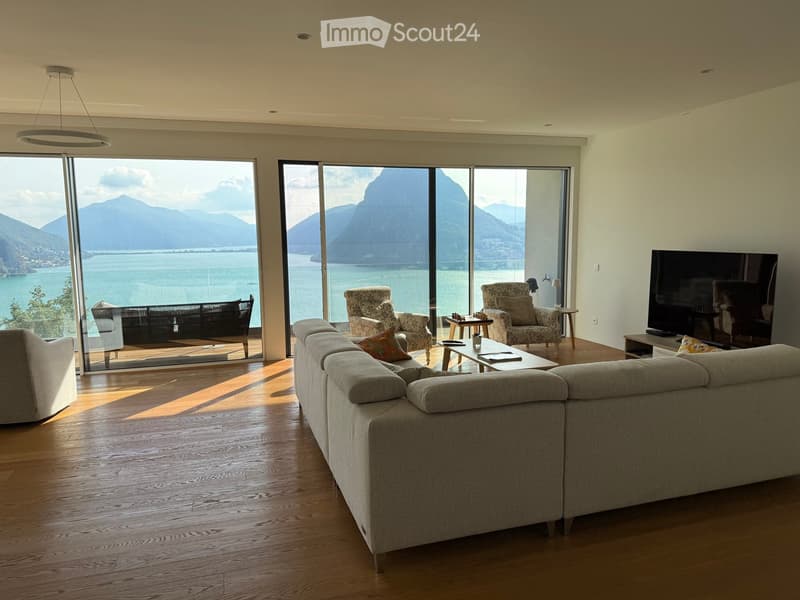 Modern living room with large windows offering a scenic mountain and lake view, featuring a grey sectional sofa, coffee table, armchairs, ottoman, and a television.