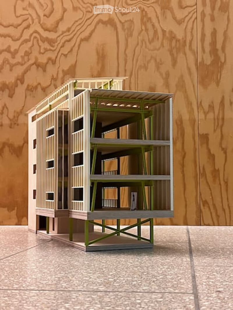 A modern, minimalist building model with a raised ground floor, green structural supports, and a combination of glass and concrete surfaces.